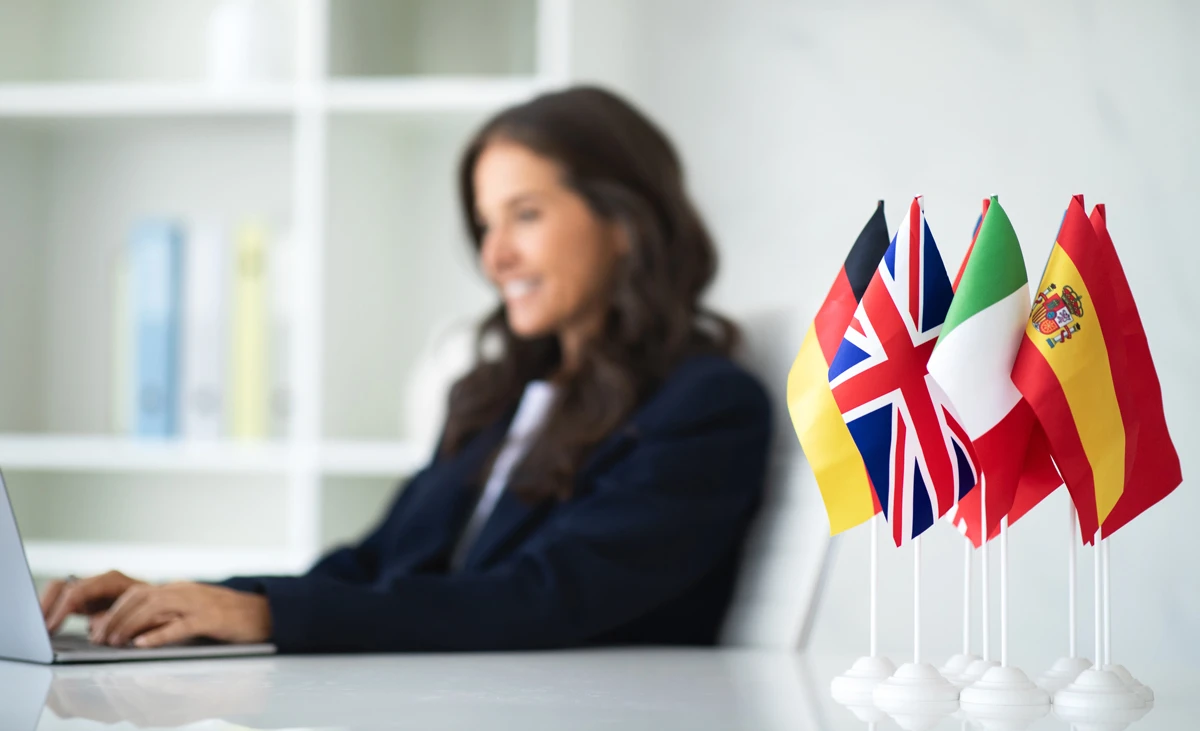 Woman working on a laptop with small UK, German, French and Spanish flags on a desk in the foreground.