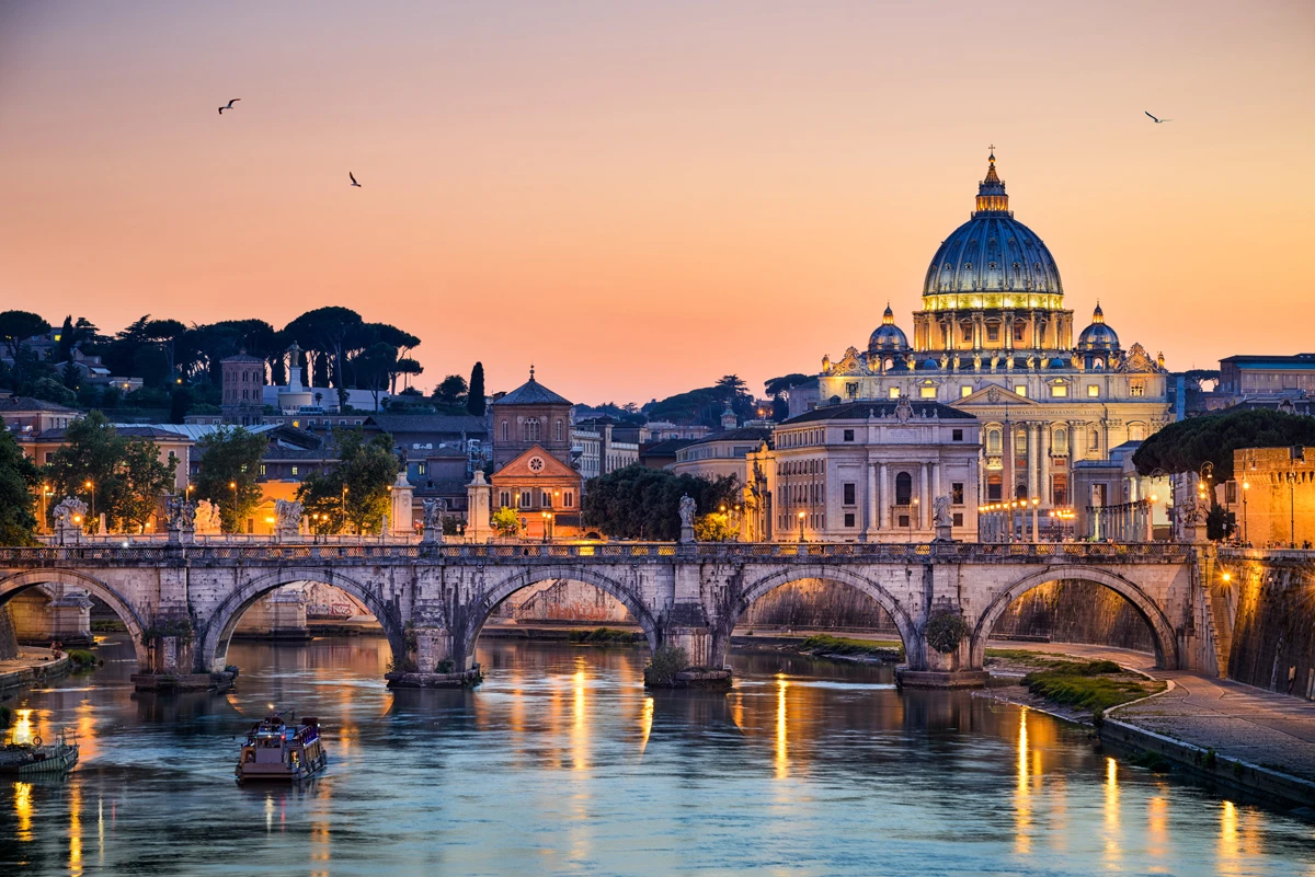 Sunset view of St Peter’s Basilica and the River Tiber in Rome, with historic bridges and warm golden lights reflecting on the water.